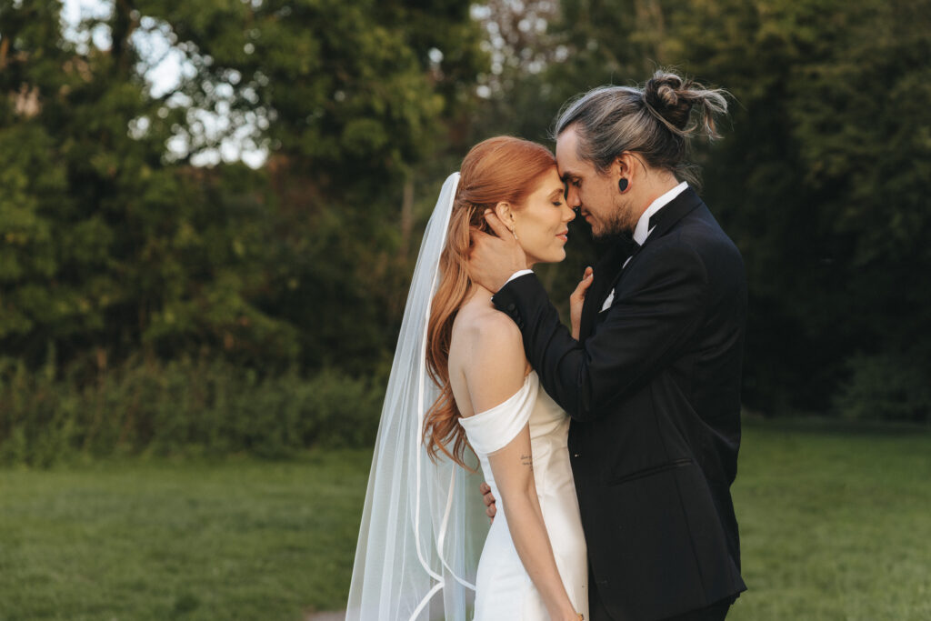 A bride with long red hair and a groom with a man-bun stand face-to-face on the grounds of The Beonna in Lincolnshire. The bride wears a white off-the-shoulder gown and veil, while the groom wears a black suit with a bow tie. © Aimee Lince Photography