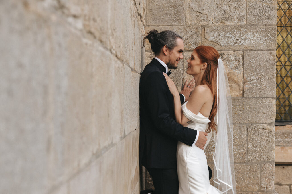 A bride in an off-the-shoulder gown and a groom in a tuxedo gaze lovingly at one another against an old stone wall at The Beonna in Lincolnshire. © Aimee Lince Photography