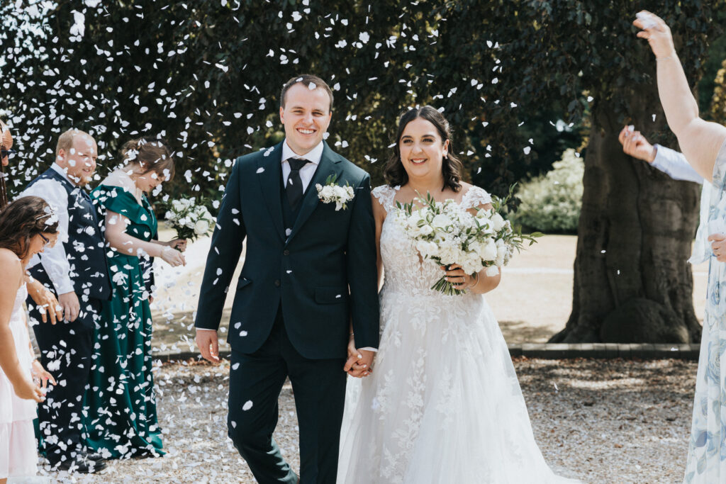 A bride and groom walk hand-in-hand outdoors as white confetti falls, captured by a wedding photographer in Lincolnshire. The groom wears a dark suit and the bride wears a white lace wedding dress with a bouquet, whilst the bridal party stands on a gravel path under trees nearby. © Aimee Lince Photography
