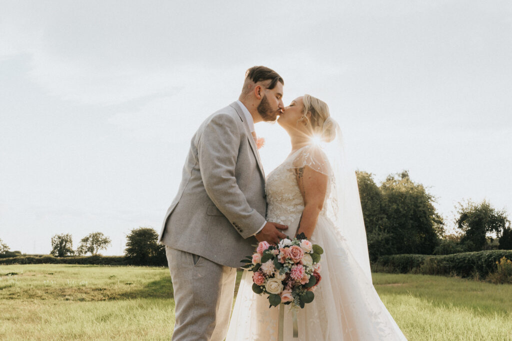 A bride and groom kiss in a grassy field during sunset, captured by Aimee Lince. The groom wears a grey suit and the bride wears a white lace wedding dress with a veil, holding a bouquet of pink and white flowers. Backlit by the sun, they stand in a Lincolnshire landscape with trees. © Aimee Lince Photography