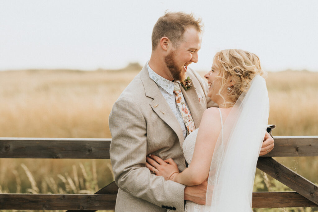 A bride in a white wedding dress and a groom in a tan suit lean towards each other, laughing by a wooden fence in a Lincolnshire field. The background is filled with tall, golden-brown grass under a soft, overcast sky, perfect for a wedding photographer. © Aimee Lince Photography