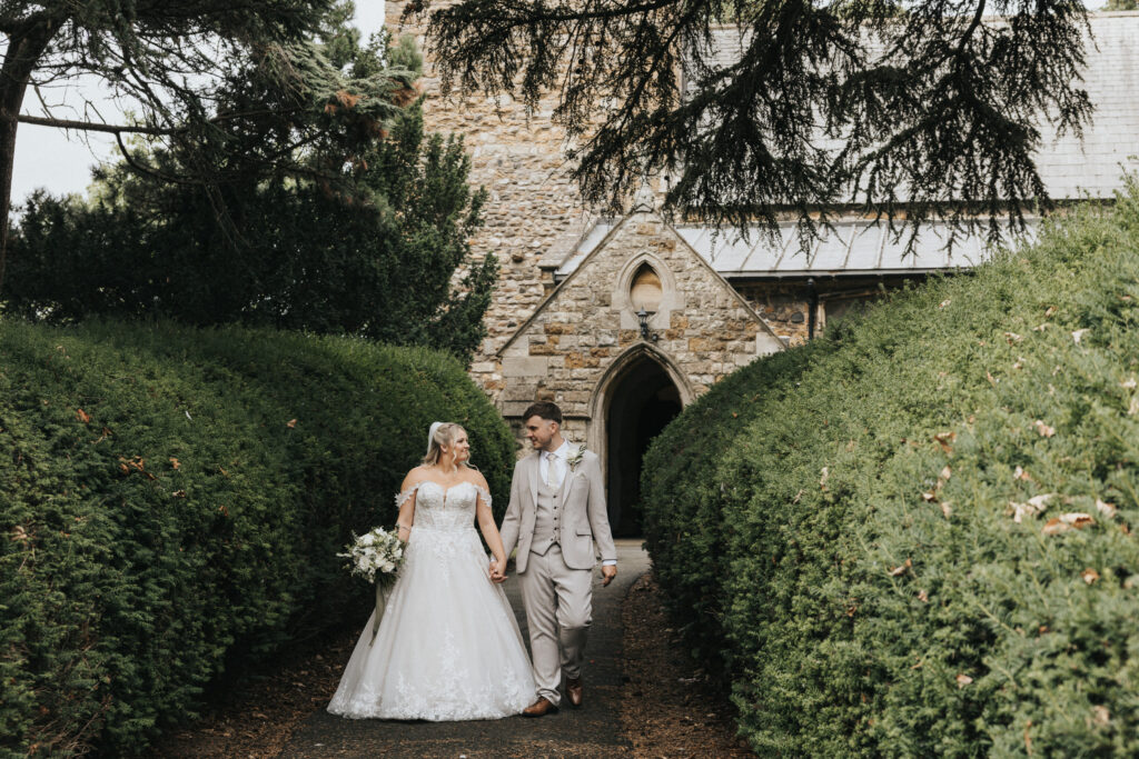 A bride in an off-the-shoulder lace gown and a groom in a tan suit walk hand-in-hand towards a stone church. Captured by a wedding photographer in Lincolnshire, the couple share a tender look whilst walking along a path framed by tall hedges, the bride holding a white bouquet. © Aimee Lince Photography