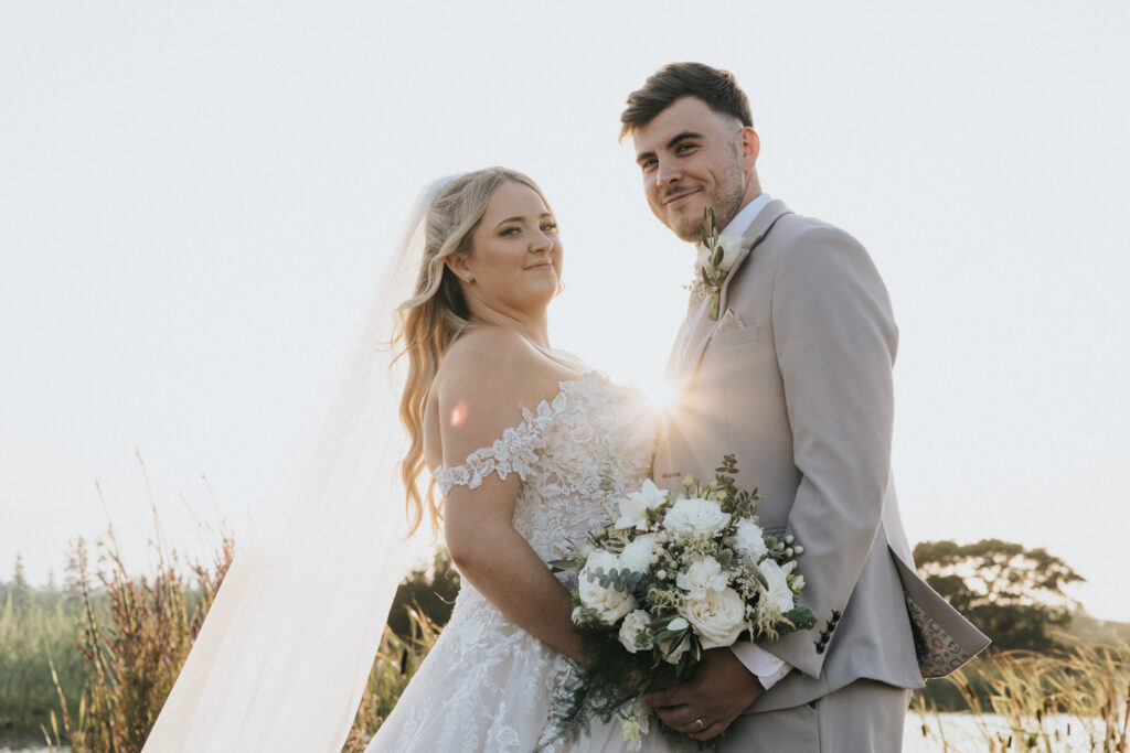 A bride and groom pose together in a sun-drenched field in this Lincolnshire wedding photograph. The bride wears a strapless lace gown with a long veil, whilst the groom wears a light-coloured suit with a buttonhole and holds a bouquet of white roses and greenery as sunlight flares between them. © Aimee Lince Photography